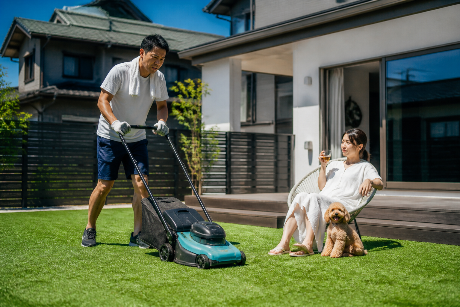 夏の炎天下で汗だくになりながら芝刈り機を押している人と、その隣で人工芝の庭に置いたチェアに座り、愛犬とくつろぎながらドリンクを飲んでいる人を対比させるイメージ写真。]